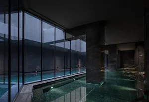 A modern indoor pool area with a large pool surrounded by a black stone wall, natural light from windows, and a black and white color scheme