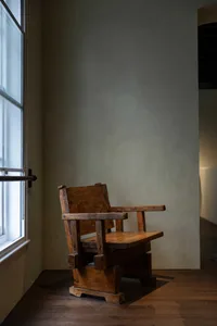 A calm and inviting room with a brown wooden chair in front of a window, surrounded by a white wall and natural light
