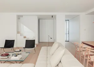A modern living room with a white couch, black chairs, and a glass coffee table, surrounded by white walls and a staircase in the background