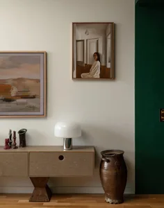 A well-decorated living room with a white wall featuring a variety of artwork, a vase on a table, and a lamp, creating an inviting atmosphere