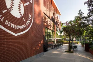 Photograph of a rustic brick building with a baseball theme, featuring a dining table and chairs in front, with a baseball glove and bat on the side, set in a casual and inviting outdoor scene