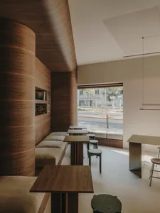A modern restaurant interior with a large wooden dining area, featuring multiple chairs, benches, and a wooden table, set against a well-lit background with a bar in the distance