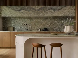 A modern kitchen with a white marble countertop, wooden stools, and a potted plant, lit by natural light