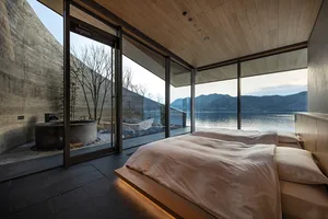 A modern hotel bedroom with a large bed, wooden headboard, and natural light from a large window