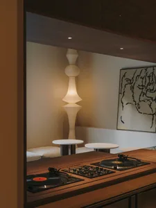 A dimly lit room with a wooden floor, a white wall, and a DJ setup with a turntable and a record player, accompanied by a couch and two chairs