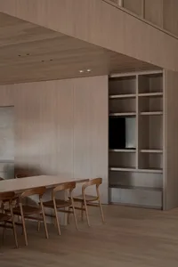 Warm and inviting living space with wooden floors and walls, a dining table, television, and bookshelf, lit by natural sunlight