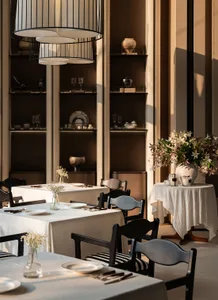 A formal dining room with a large table set with white tablecloths, wine glasses, cups, and bowls, surrounded by comfortable chairs and decorative elements like vases and potted plants, with soft and warm lighting creating a cozy ambiance