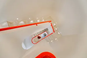 A modern staircase in a white room with red handrails and a potted plant on the stairs, set in a dimly lit space