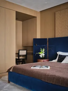 Cozy bedroom with a blue-covered bed, a wooden floor, and a brown color scheme, with a vase and flowers on a table, and a book on the bed, in a dimly lit space