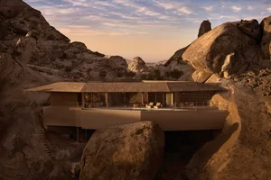 A modern house with a glass facade and concrete construction, situated in front of a large rock in a mountainous landscape, with a bright and inviting interior