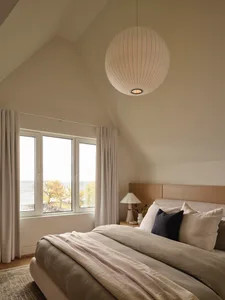 A calm bedroom with a white bed, a window, and a table lamp, surrounded by a white color scheme and natural light