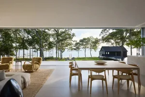 A well-lit living room with a dining table, couch, and chair, surrounded by a large window showcasing a beautiful water view