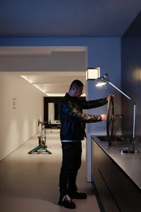 A man in a bathroom, standing next to a sink and a bicycle, with a toilet in the background, wearing a leather jacket and adjusting a bike wheel, in a white-colored room with a light on the wall