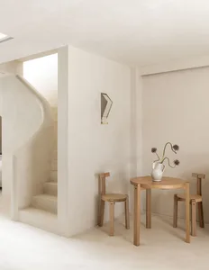 A bright and welcoming living room with a white color scheme, a staircase, dining table, and vase with flowers, lit by natural sunlight through the windows