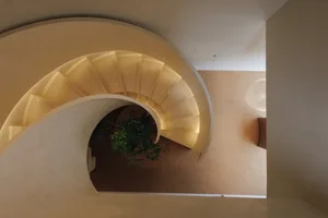 A spiral staircase in a dimly lit room with a white wall, made of wood with a brown color