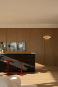 Modern kitchen with wooden walls and cabinets, featuring a counter with a sink, refrigerator, and oven, with two red stools and a potted plant, lit by natural sunlight
