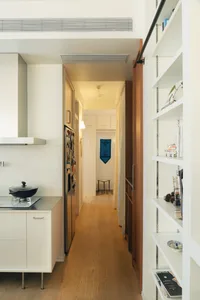 A predominantly white kitchen and hallway with a refrigerator, sink, and bowl on the counter, a doorway, and a flag on the wall, lit by natural light