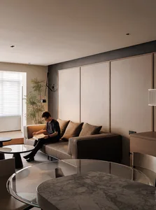 Cozy living room with a man reading on the couch, surrounded by a dining table, chair, and potted plant, with a warm and inviting atmosphere