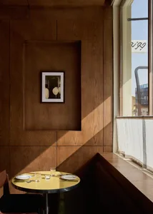A modern dining space with a wooden table, chairs, and bench, set with plates, cups, and a wine glass, surrounded by a wooden wall and natural light