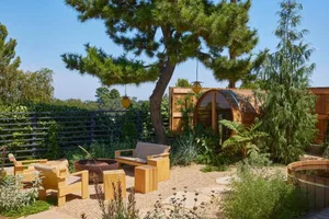 A serene outdoor patio with a wooden deck, gazebo, and natural surroundings, featuring a wooden bench and chair on the dirt ground