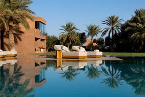 A modern poolside patio with a lounge chair surrounded by palm trees and a clear blue pool