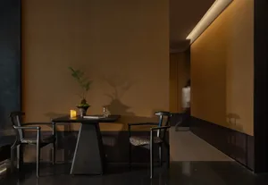 Dimly lit dining room with a wooden floor, a brown table, and black chairs, surrounded by a potted plant