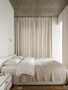A calm bedroom with a white bed, nightstand, and curtains, lit by natural light from the window