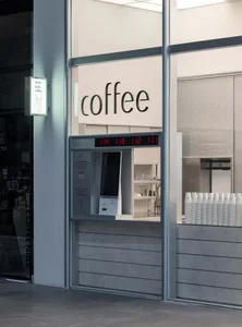 A modern coffee shop with a glass front, white walls, and a sleek metal counter, featuring a coffee machine, cups, and bottles, under bright and welcoming lighting