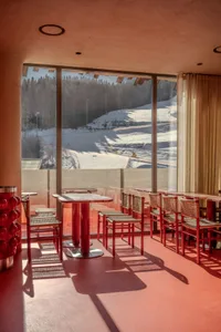 Cozy restaurant with a large dining table and red chairs, surrounded by a modern space with a large window offering a view of snow-covered mountains