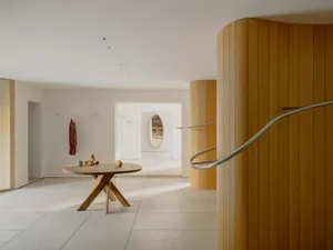 A warm and cozy room with a wooden floor, white wall, and wooden staircase and table, featuring a mix of natural and artificial lighting