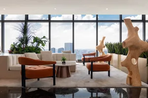A modern living room with a large window showcasing a city view, featuring a couch, chair, and dining table, with potted plants and natural light creating a bright and inviting atmosphere