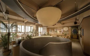 A modern dining space with a circular light fixture, multiple chairs, and dining tables, surrounded by a large window allowing natural light to fill the space
