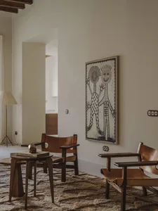 Comfortable living space with a white wall, a large painting, a dining table, a couch, and a lamp, well-lit by natural light from a window