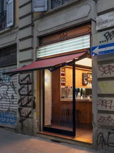 A modern bar with a red awning, graffiti-covered walls, a bar with a refrigerator and sink, and bottles on the counter, set in a dimly lit storefront