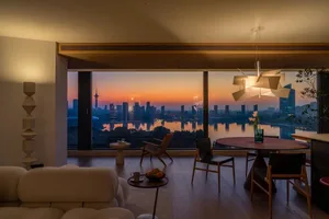 A modern living room with a large window overlooking the city, featuring a couch, dining table, chair, vase, potted plant, and various decorative items, with a mix of natural and artificial lighting