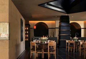 A modern restaurant with a wooden dining table and chairs, set with various items, surrounded by natural light from the windows