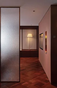 A modern hallway with a pink wall, wooden floor, and a couch in the middle, lit by two lamps and a mirror on the right side