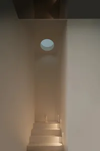 A dimly lit white room with a small statue on a pedestal and a round window, creating a mysterious atmosphere