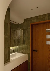 Bathroom with a wooden door and green tile wall, featuring a sink and bathtub in a modern space with dim lighting