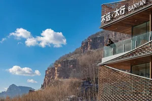 一名男子站在一塊玻璃陽台上，俯瞰著一座山脈風光的風景，天空是靜穩而藍藍的，自然光線照射下