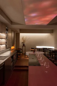 A modern restaurant with a long dining table, wooden chairs, and wine glasses, set under a light fixture, creating a sophisticated ambiance