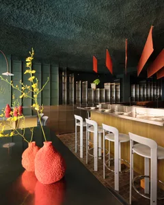 A modern kitchen with a black counter, white stools, and a red vase, lit by natural light
