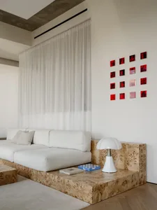 A modern living room with a white couch, chair, and lamp, surrounded by red squares on the wall, with natural light and a calm atmosphere