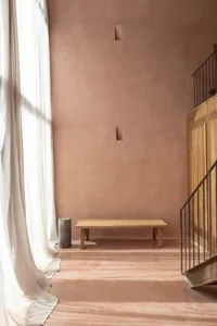 A cozy living room with a wooden staircase, a wooden bench, and a pink wall, lit by natural sunlight