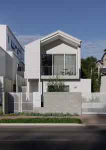 A modern beach house with white walls, a brick wall, and a fence, featuring a balcony, dining table, and potted plant, with natural light and a welcoming atmosphere