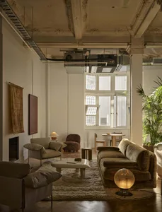 A modern living room with a white color scheme, a large couch, a chair, and a dining table, lit by natural sunlight