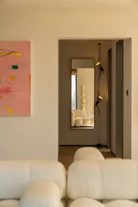 A well-lit living room with a white couch and a chair, decorated with a pink painting and a mirror, creating a comfortable atmosphere