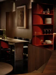 A modern dining room with a wooden table and chairs, surrounded by a red bookshelf, in a dimly lit space
