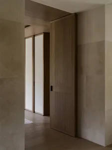 A small room with a wooden door and a white wall, featuring a wooden floor and a doorway leading to another room, with dim lighting
