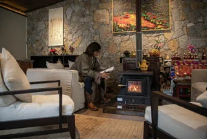 Cozy living room with a stone wall, fireplace, and natural materials, featuring a man sitting on a couch surrounded by chairs, a potted plant, and a vase